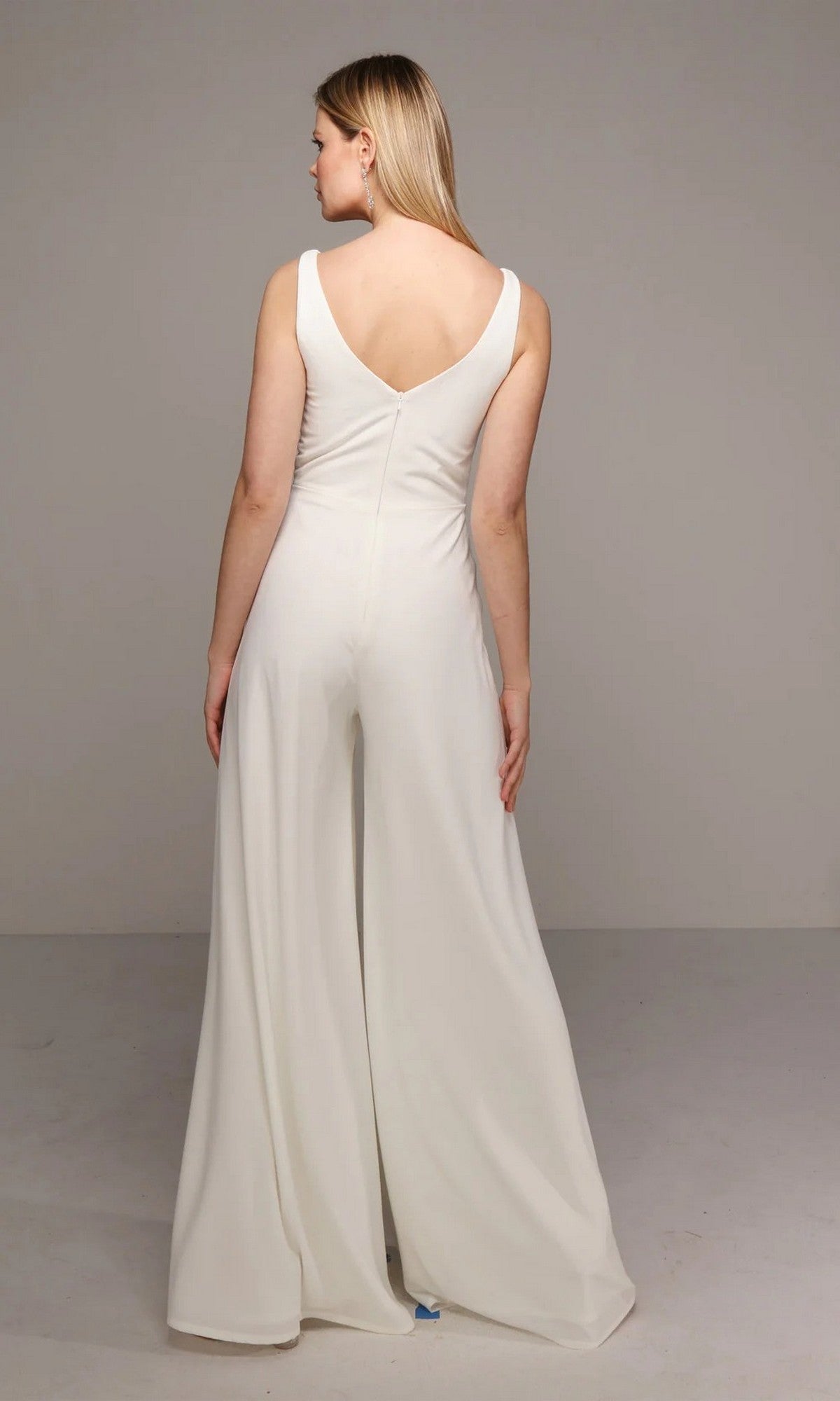 A woman faces away, wearing the Alyce 70021 long white jumpsuit with a sleeveless design, wide-leg pants, and a V-shaped back, set against a plain gray background.
